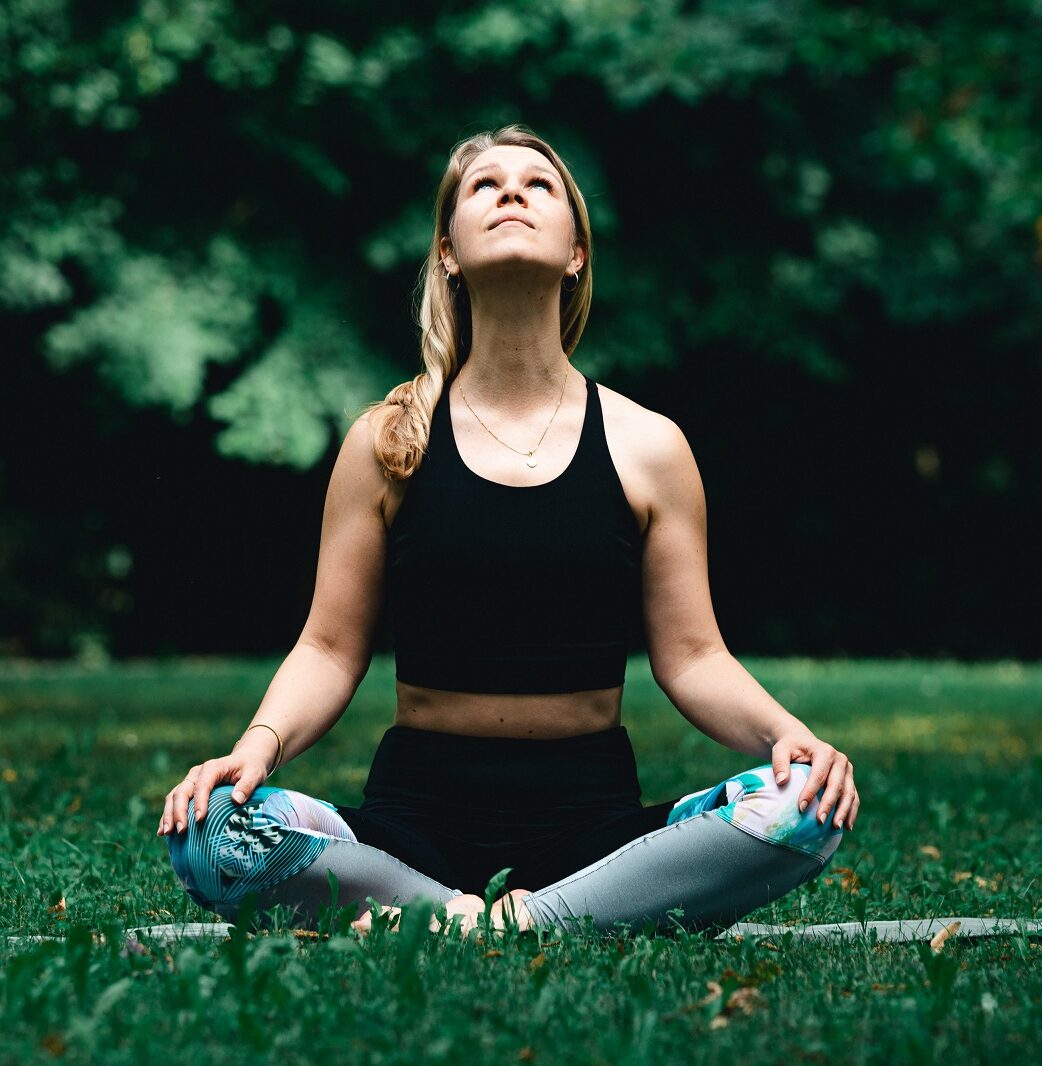 Eine Frau sitzt im Schneidersitz, die Hände entspannt auf den Knien abgelegt, und blickt zum Himmel hoch.